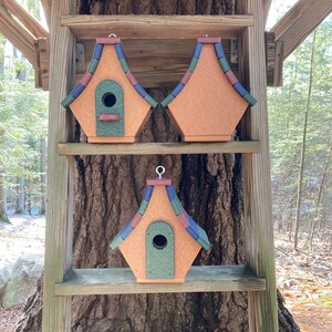 Op de afbeelding: Drie vogelhuisjes met kleurrijke, geschindeld daken worden getoond op een houten plank. De vogelhuisjes zijn gemaakt van gerecycled plastic en hebben een groen, bruin en blauw kleurenschema.