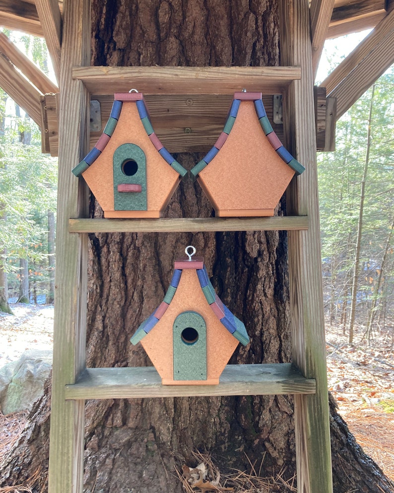 Op de afbeelding: Drie vogelhuisjes met kleurrijke, geschindeld daken worden getoond op een houten plank. De vogelhuisjes zijn gemaakt van gerecycled plastic en hebben een groen, bruin en blauw kleurenschema.