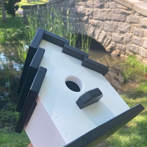 May include: A white birdhouse with black trim. The birdhouse has a round entrance hole and a small perch on the front. The birdhouse is sitting on a hand.