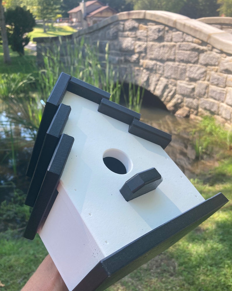 May include: A white birdhouse with black trim. The birdhouse has a round entrance hole and a small perch on the front. The birdhouse is sitting on a hand.