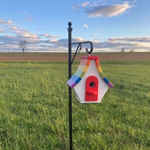 May include: A rainbow-colored birdhouse with a red entrance hole, mounted on a black metal pole against a green grassy field.