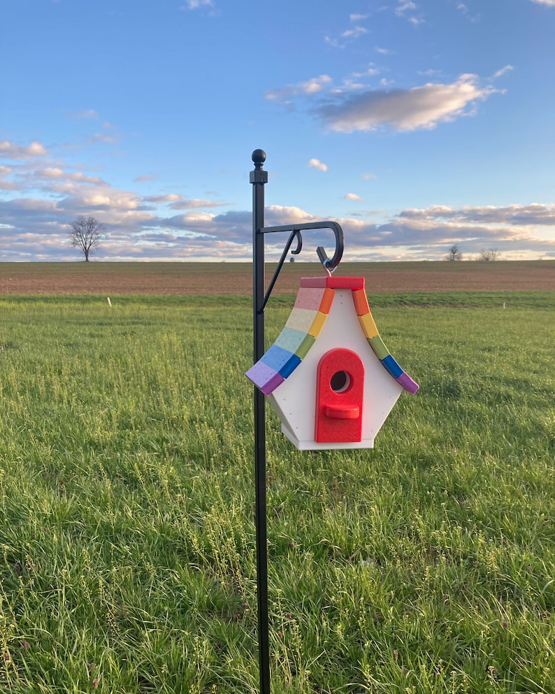 May include: A rainbow-colored birdhouse with a red entrance hole, mounted on a black metal pole against a green grassy field.