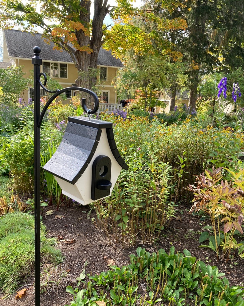 May include: A white and black birdhouse with a black roof hangs from a black metal stand in a garden setting. The birdhouse has a small opening for birds to enter.