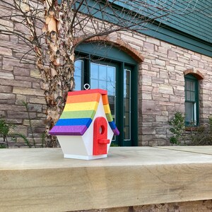 May include: A rainbow-colored birdhouse with a red door. The birdhouse is made of wood and has a white base. It is sitting on a stone ledge in front of a brick building.