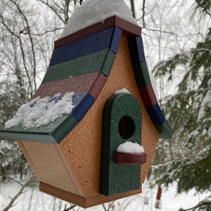 Op de afbeelding: Een kleurrijk houten vogelhuisje met een groen, blauw en rood gestreept dak. Het vogelhuisje is bedekt met sneeuw en heeft een kleine opening waar vogels naar binnen kunnen.