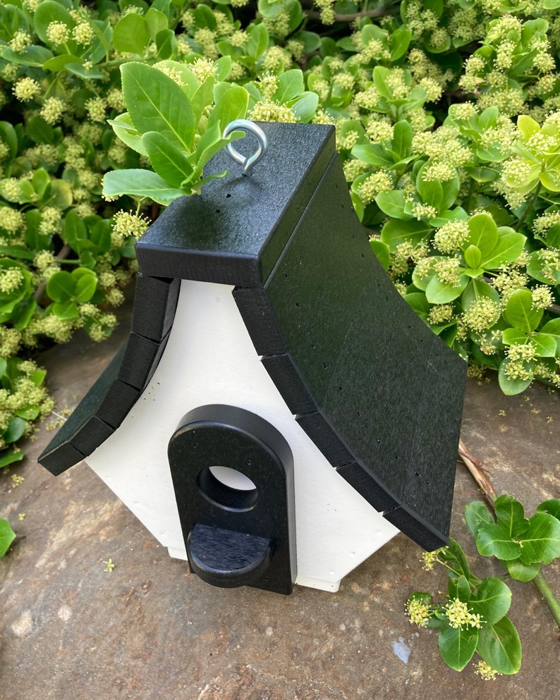 May include: A black and white birdhouse with a peaked roof and a small entrance hole. The birdhouse has a metal hook for hanging.