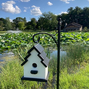 May include: A white birdhouse with a black roof hangs from a black metal pole. The birdhouse has a round opening for birds to enter. The birdhouse is in front of a pond with green lily pads and a blue sky with white clouds.