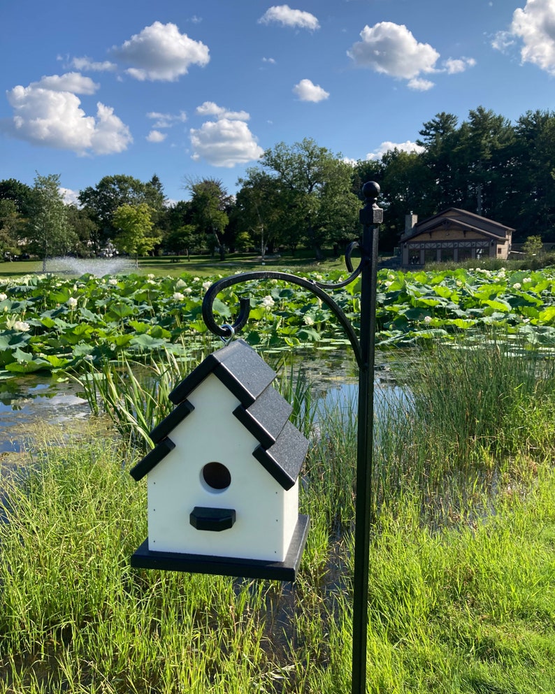 May include: A white birdhouse with a black roof hangs from a black metal pole. The birdhouse has a round opening for birds to enter. The birdhouse is in front of a pond with green lily pads and a blue sky with white clouds.