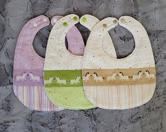 May include: Three baby bibs with a zebra print design. The bibs are made of a soft, white fabric with a green, purple, and brown trim. Each bib has a different colour trim and a different pattern of zebras.