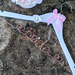 May include: White wooden hanger with a pink bow and a white flower. The word "Bride" is written in copper wire.