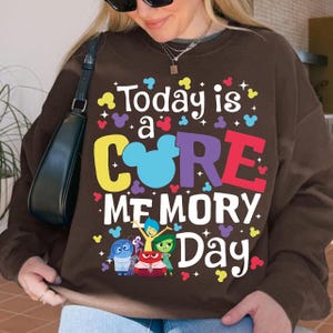 May include: A brown sweatshirt featuring the phrase "Today is a CORE MEMORY Day" in vibrant colours. The design incorporates Mickey Mouse ears and cartoon characters. The sweatshirt is a relaxed fit.