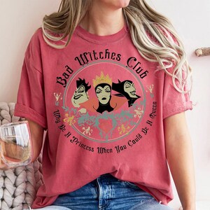 Peut inclure: T-shirt rouge avec un motif circulaire repr&eacute;sentant trois personnages de sorci&egrave;res de dessins anim&eacute;s et le texte "Bad Witches Club". Le motif comprend la phrase "Why Be A Princess When You Could Be A Queen."