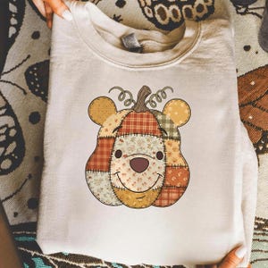 May include: A cream-colored sweatshirt featuring a patchwork pumpkin design with a bear face. The pumpkin has a brown stem and is made of various patterned fabrics in shades of orange, brown, and yellow. The bear face has a smiling expression.