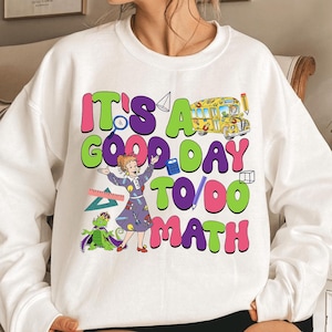 May include: White sweatshirt with a colorful graphic that reads "It's a Good Day to Do Math." The design includes a school bus, a teacher, a dragon, and math-related symbols in bright colors.
