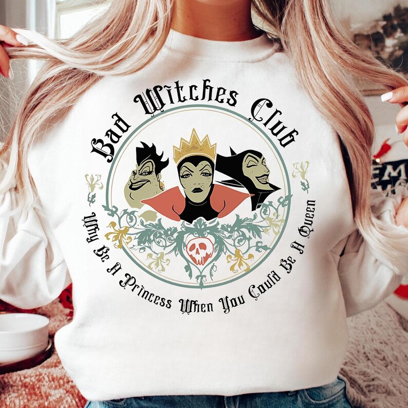 Peut inclure: Sweat-shirt blanc avec un motif circulaire. Le motif comprend le texte "Bad Witches Club" et "Why Be A Princess When You Could Be A Queen". Le motif comprend trois sorci&egrave;res de dessin anim&eacute; en teal, or et rouge, avec des accents floraux.