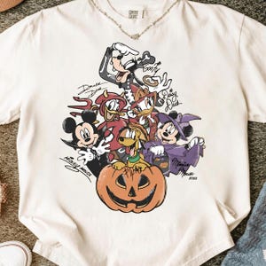 Op de afbeelding: Crèmekleurig T-shirt met een Halloween-thema van Disney-personages, waaronder Mickey Mouse, Donald Duck en Goofy, in kostuums. Het ontwerp bevat een uitgesneden pompoen en handtekeningen van personages.