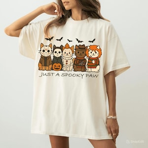 Op de afbeelding: Crèmekleurig T-shirt met een Halloween-thema. Het ontwerp bevat cartoonkatten verkleed als horrorfilmkarakters, vleermuizen en de tekst "JUST A SPOOKY PAW".