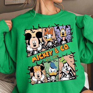 May include: Green sweatshirt featuring a Halloween-themed graphic of Mickey Mouse, Minnie Mouse, Donald Duck, Daisy Duck, and Goofy in skeleton form. The text "MICKEY & CO. EST. 1928" is printed in orange. Black bat silhouettes are scattered around the design.