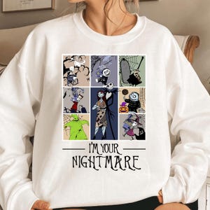 Puede incluir: Sudadera blanca con una cuadrícula de personajes de dibujos animados de "Pesadilla antes de Navidad". El diseño incluye a Jack Skellington, Sally y otros personajes. El texto "I'M YOUR NIGHTMARE" está impreso en negro debajo.