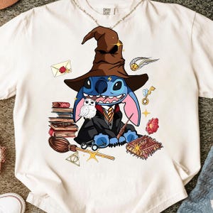 May include: White t-shirt featuring a cartoon Stitch character dressed as a wizard, wearing a brown hat and black robe. The design includes books, a broom, a golden snitch, and other magical elements.