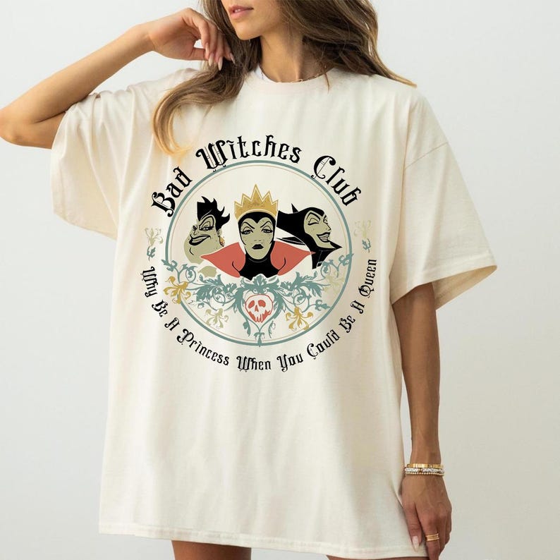 Peut inclure: T-shirt cr&egrave;me avec un motif circulaire. Le motif affiche le texte "Bad Witches Club" et pr&eacute;sente trois personnages de m&eacute;chants, une couronne et la phrase "Why Be A Princess When You Could Be A Queen."