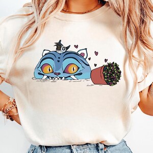 May include: Cream-colored t-shirt featuring a whimsical illustration. A blue cat-like creature with yellow eyes and sharp teeth is depicted, along with a small bird wearing a hat. A potted plant with purple berries and hearts complete the design.
