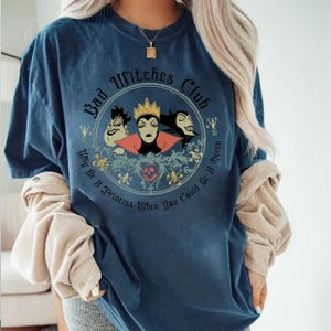 Peut inclure: T-shirt bleu marine avec un motif circulaire. Le motif comprend le texte "Bad Witches Club" et "Why Be A Princess When You Could Be A Queen", ainsi que des illustrations de trois sorci&egrave;res de dessins anim&eacute;s.