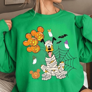 May include: Green sweatshirt featuring a Halloween-themed design. The graphic includes a mummy-wrapped dog, Mickey Mouse pumpkin balloons, ghosts, bats, and a spiderweb. The design is in orange, black, white, and yellow.