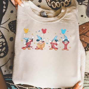 Könnte beinhalten: Beiges Sweatshirt mit einem farbenfrohen Aufdruck von Mickey Mouse in einem Zauberer-Outfit. Mickey wird mit gelben, roten und blauen Luftballons dargestellt. Das Design enthält Sterne und Herzen und erzeugt eine skurrile und verspielte Ästhetik.