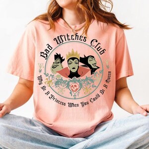 Peut inclure: T-shirt p&ecirc;che avec un motif circulaire. Le motif comprend le texte "Bad Witches Club" et des illustrations de trois sorci&egrave;res de dessins anim&eacute;s. La phrase "Why Be A Princess When You Could Be A Queen" est &eacute;galement incluse.