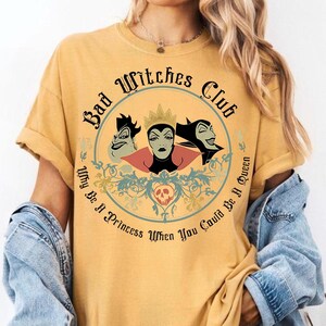 Peut inclure: T-shirt jaune moutarde avec un motif circulaire. Le motif comprend le texte "Bad Witches Club" et "Why Be A Princess When You Could Be A Queen", ainsi que des illustrations de trois sorci&egrave;res de dessins anim&eacute;s.