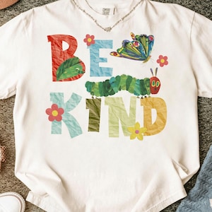 以下が含まれることがあります： カラフルな文字で「BE KIND」と書かれた白いTシャツ。デザインには、緑色のイモムシ、蝶、花が含まれています。柔らかい素材で作られており、クルーネックです。