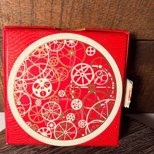 May include: A red square box with a circular design of gold and white gears. The box has a textured surface and a small piece of paper attached to the side. The gears are of various sizes and styles.