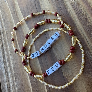 Op de afbeelding: Drie armbanden met kralen met de tekst "GO NOLES FSU". De armbanden zijn gemaakt met gouden, rode en transparante kralen. De armbanden liggen op een houten oppervlak.
