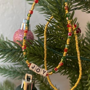Pode incluir: Um colar de contas douradas com contas vermelhas e verdes, e um amuleto que diz "JOY" pendurado nele. O colar est&aacute; pendurado em uma &aacute;rvore de Natal.