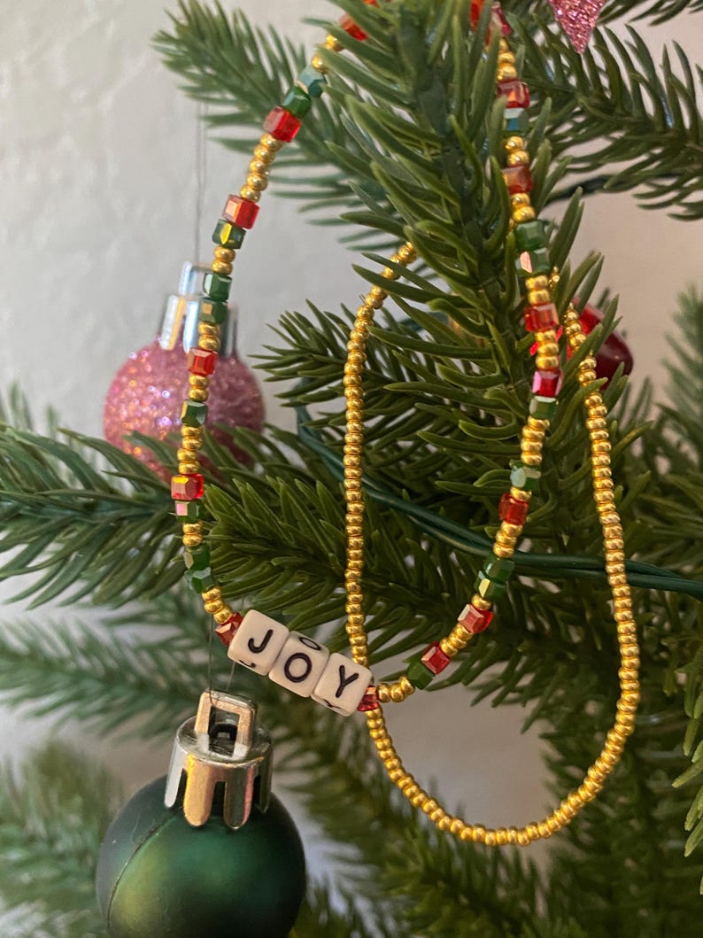 Pode incluir: Um colar de contas douradas com contas vermelhas e verdes, e um amuleto que diz "JOY" pendurado nele. O colar est&aacute; pendurado em uma &aacute;rvore de Natal.