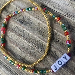 Pode incluir: Um cord&atilde;o de contas de cintura dourado com contas vermelhas, verdes e iridescentes. O cord&atilde;o de contas tem um amuleto branco com a palavra "JOY" nele.