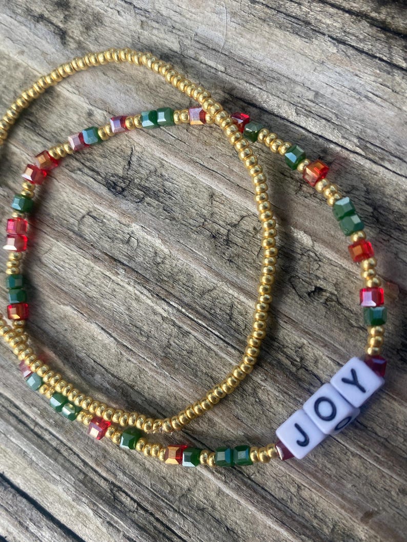 Pode incluir: Um cord&atilde;o de contas de cintura dourado com contas vermelhas, verdes e iridescentes. O cord&atilde;o de contas tem um amuleto branco com a palavra "JOY" nele.