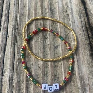 Pode incluir: Duas pulseiras de contas com contas douradas, vermelhas e verdes. A pulseira menor tem a palavra "JOY" escrita em contas de letras brancas.