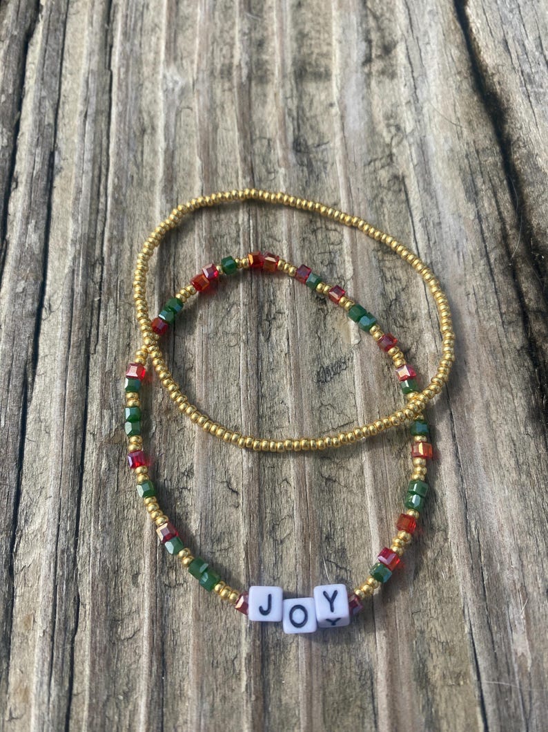 Pode incluir: Duas pulseiras de contas com contas douradas, vermelhas e verdes. A pulseira menor tem a palavra "JOY" escrita em contas de letras brancas.