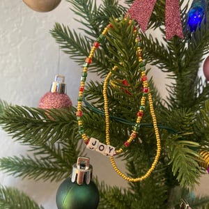 Pode incluir: Um enfeite de &aacute;rvore de Natal com um colar de contas e a palavra "JOY" escrita em contas brancas.