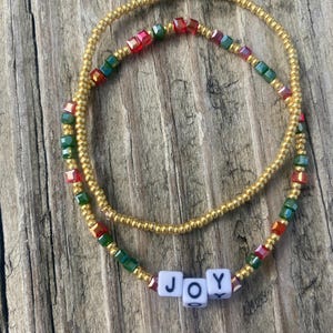 Pode incluir: Duas pulseiras de contas douradas com contas vermelhas e verdes iridescentes. As pulseiras s&atilde;o sobrepostas e soletram a palavra "ALEGRIA" em pe&ccedil;as de letras brancas.