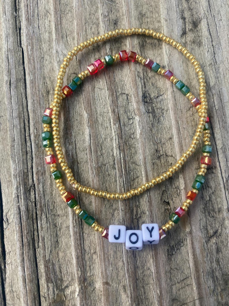 Pode incluir: Duas pulseiras de contas douradas com contas vermelhas e verdes iridescentes. As pulseiras s&atilde;o sobrepostas e soletram a palavra "ALEGRIA" em pe&ccedil;as de letras brancas.