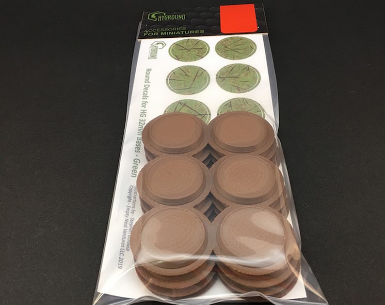 May include: A package of miniature accessories. The package contains several brown, circular bases and a sheet of green decals. The text on the package reads "SHYGROUND ACCESSORIES FOR MINIATURES" and "Round Decals for HG 32mm Bases - Green".