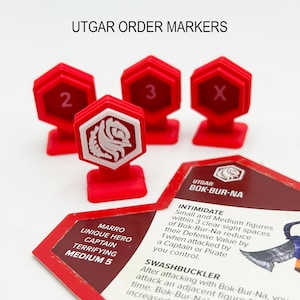 Könnte beinhalten: Rote Plastik-Ordnungsmarker mit den Zahlen 2, 3 und X. Ein roter Plastikmarker mit einem weißen Drachenmotiv. Eine rote Karte mit dem Text "UTGAR BOK-BUR-NA" und dem Text "INTIMIDATE" und "SWASHBUCKLER".