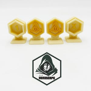 Könnte beinhalten: Vier gelbe, sechseckige Spielsteine aus Plastik mit verschiedenen Symbolen. Die Spielsteine liegen auf einer weißen Oberfläche. Die Symbole sind eine 1, eine Person mit Kapuze, eine Person mit Kapuze und ein X.