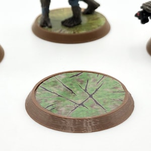 May include: A brown, circular miniature base with a textured, green and brown surface. The base is designed for tabletop gaming miniatures, providing a detailed and thematic foundation for figures. The base is approximately 1 inch in diameter.