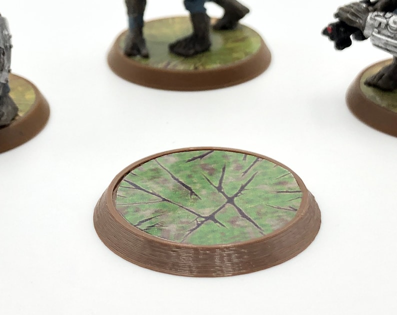 May include: A brown, circular miniature base with a textured, green and brown surface. The base is designed for tabletop gaming miniatures, providing a detailed and thematic foundation for figures. The base is approximately 1 inch in diameter.