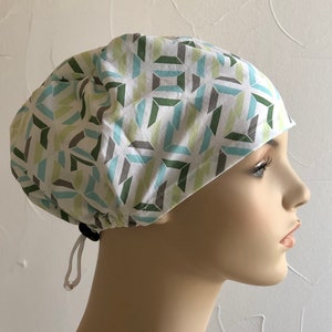 May include: A white scrub cap with a geometric pattern in shades of green, blue, and gray. The cap has a drawstring closure.