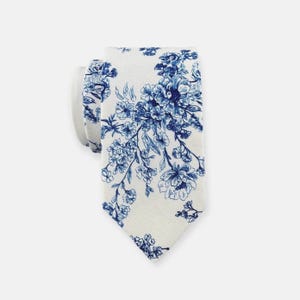 Op de afbeelding: Een witte stropdas met een blauw bloemenpatroon. De stropdas is gemaakt van een zachte stof en heeft een klassieke vorm. Het bloemmotief is gedetailleerd en bedekt de hele stropdas, wat een stijlvol accessoire creëert.